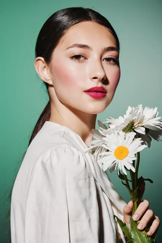 Young woman with glossy dark hair wearing bright lipstick holds white daisies against green background. Studio shot highlights natural fresh beauty and fashion creative concept.