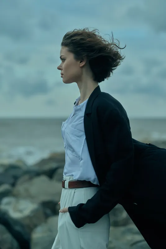 Woman walking along a windy coastline in a blazer and white blouse, carrying a bag, contemplating the sea breeze, fashion mood, urban elegance meeting coastal landscape