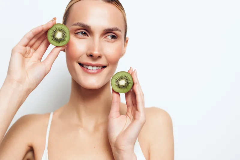 A smiling young woman holds two halves of fresh kiwi fruit near her face against a white background. Concept for healthy lifestyle, natural beauty, and fresh fruit consumption.