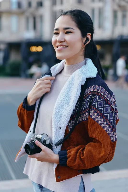 Smiling woman outdoors with camera, casual street jacket, urban setting, candid photography for lifestyle storytelling and travel vibe