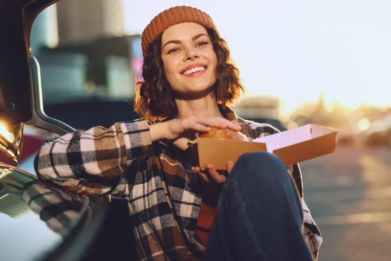 woman smile food car golden candid lifestyle relaxed authenticity enjoying a takeaway box by open trunk at sunset with golden hour glow, mindful living and emotional storytelling vibe
