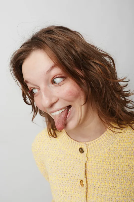 Playful young woman with short brown hair and freckles sticks out her tongue while looking sideways. She wears a yellow knitted sweater against a plain light background. Fun and carefree mood.