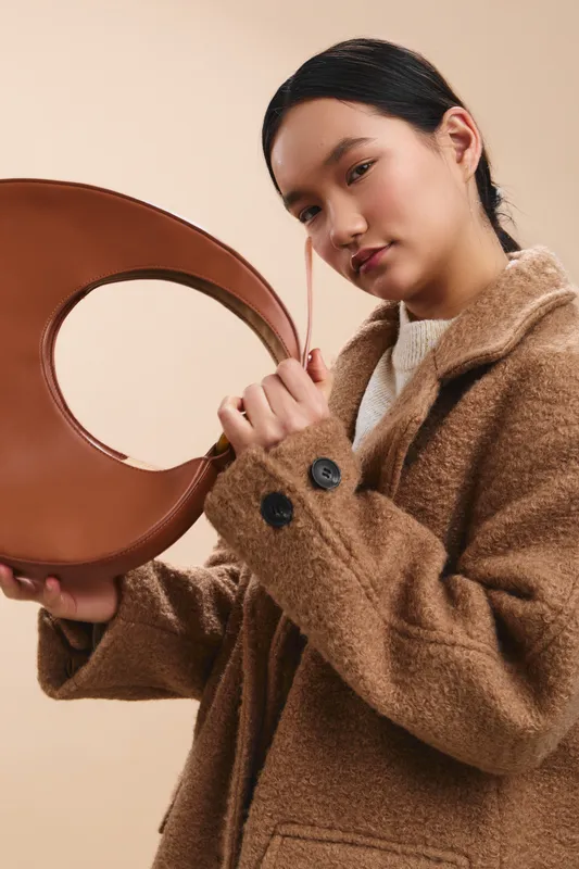 asian woman wearing a camel coat holds a round leather belt, posing against a warm neutral backdrop. she exudes chic style and calm confidence, inviting curiosity about fashion.