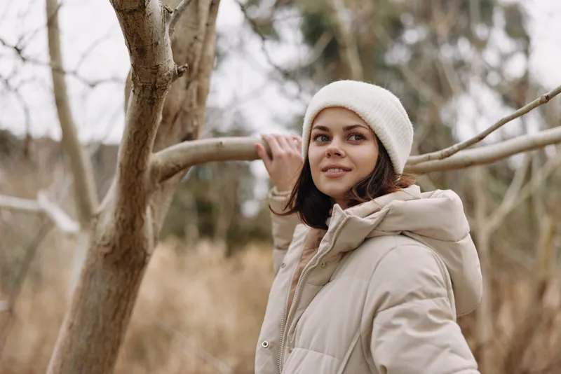 Woman portrait in a winter outdoor setting, wearing a beige coat and beanie, standing among bare trees with soft natural light and a calm, earthy mood.