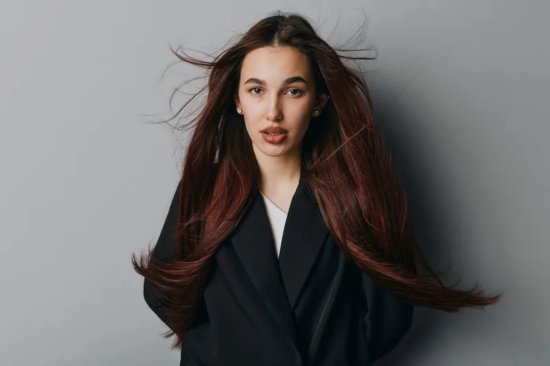 Young woman with long brown hair wearing black blazer posing on gray background. Portrait of confident female model with natural makeup and flowing hair in studio for fashion or beauty.