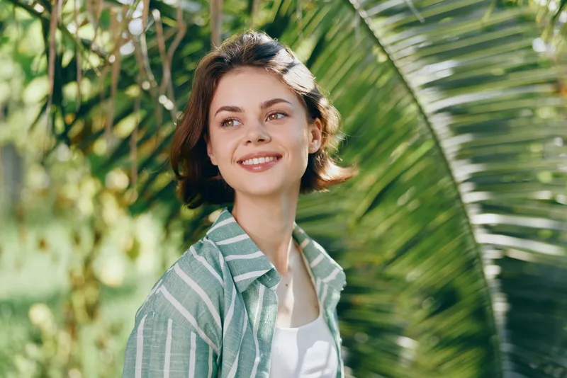 Smiling young woman outdoors among tropical greenery, wearing a striped green shirt and white top.