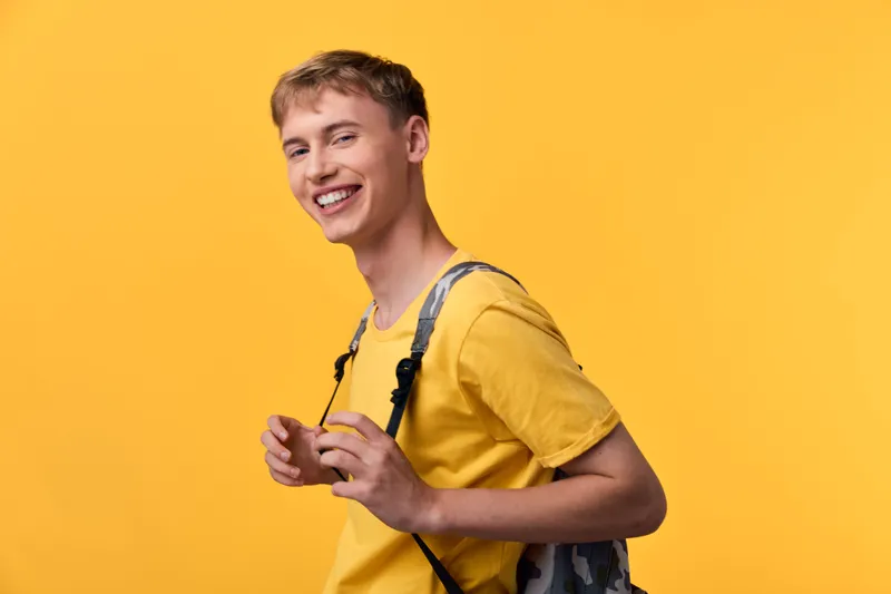 Cheerful man wearing a yellow shirt with a backpack posing in a bright studio for lifestyle and fashion imagery, capturing energy, confidence, casual style, and a welcoming mood