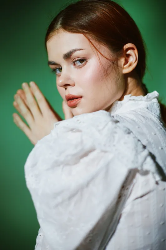 A close up portrait of a young woman with fair skin and long hair styled elegantly, wearing a vintage white blouse, exuding a calm and thoughtful expression against a green background