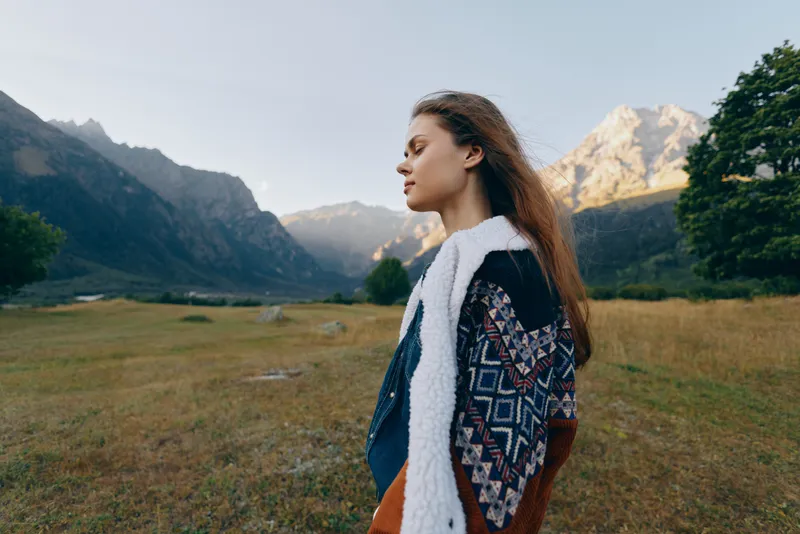 Woman in profile stands in mountain meadow wearing patterned jacket, nature landscape and golden sunlight. Young portrait outdoors, serene hiking scene and peaceful alpine scenery.