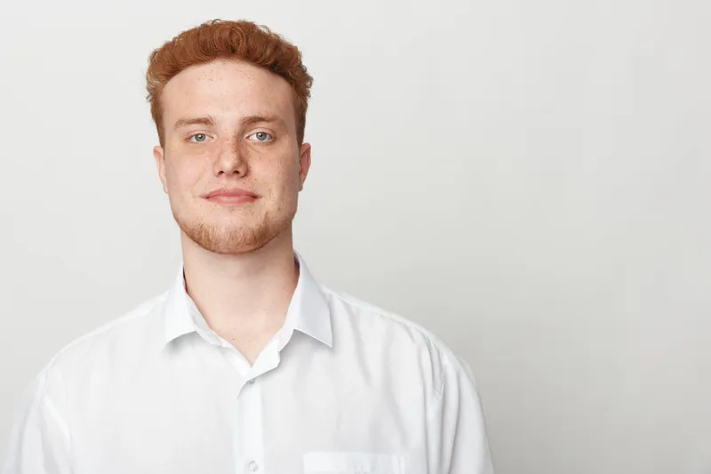 Portrait of a confident young man with red hair wearing a white shirt standing against a plain light background. Concept of casual style, youth, and natural expression in studio lighting.