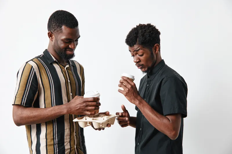 Two young black men with dark skin holding coffee cups and a cardboard tray, standing and smiling against a white background in casual clothing and short hair styles.