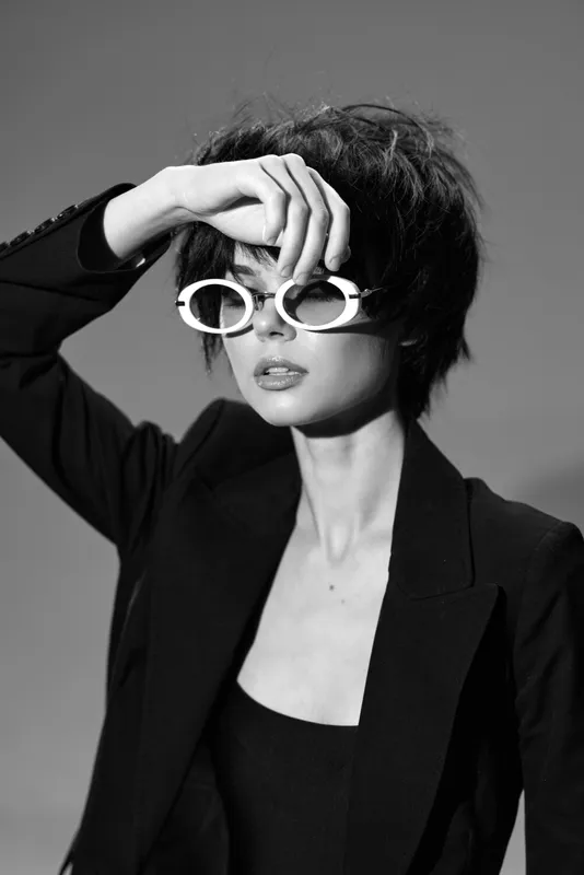 Black and white portrait of stylish woman wearing round glasses and dark blazer with short hair, posing with raised hand