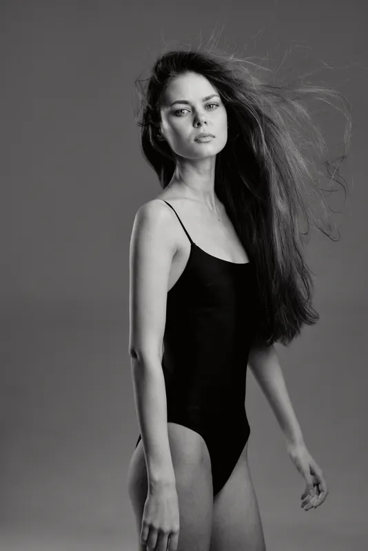 black and white portrait of a young woman with long hair in a black bodysuit standing against gray background