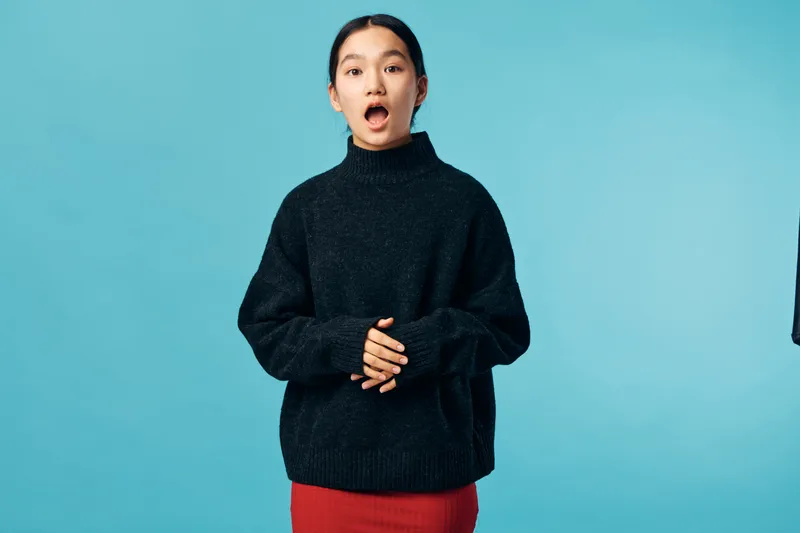 Young woman with dark hair in black sweater and red skirt standing against blue background looking surprised with open mouth and clasped hands in front.