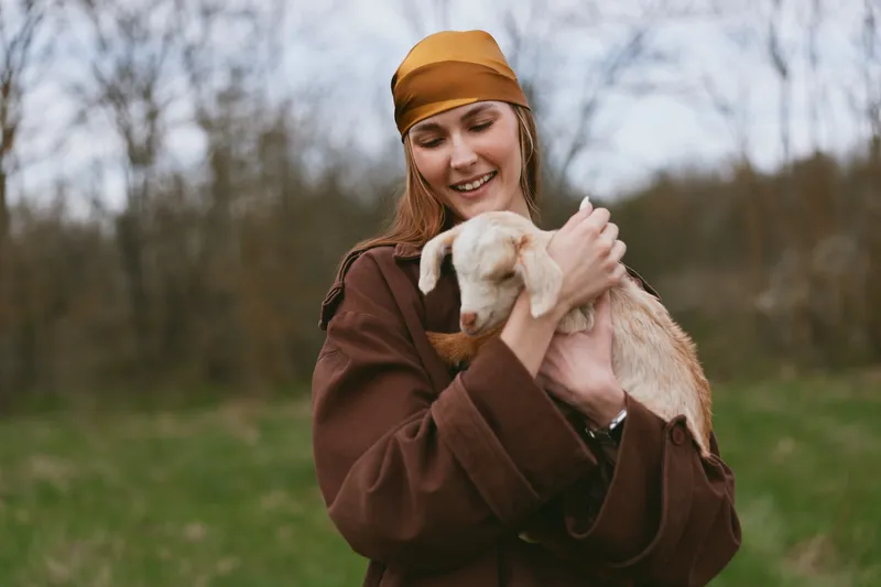 Young woman in a brown coat and amber headband smiles while gently holding a playful goat kid in an open field, capturing a warm lifestyle moment outdoors.