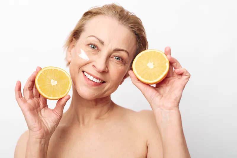 Smiling mature woman holds two halves of a grapefruit near face with natural skin cream on cheek. Concept of healthy lifestyle, skincare routine, and natural beauty on white background.