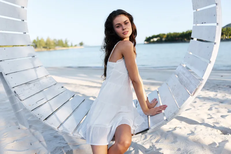 Young woman in a white sundress sits on a circular white swing on a sunny beach, relaxed pose, calm sea behind, warm natural light, summery mood and freedom.
