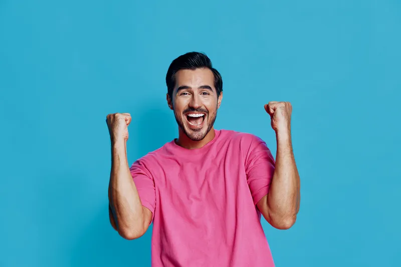 Full body excited happy cheerful young man wearing pink t-shirt celebrating success with raised fists smiling at camera isolated on solid blue background.