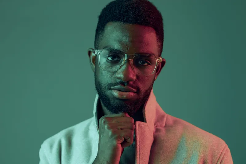 A confident young man with glasses poses against a green background, showcasing a modern style and strong emotions of determination