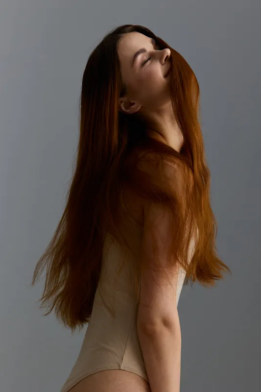 Young woman with long red hair wearing beige bodysuit posing gracefully against gray background in soft natural light, evoking calm and elegance.
