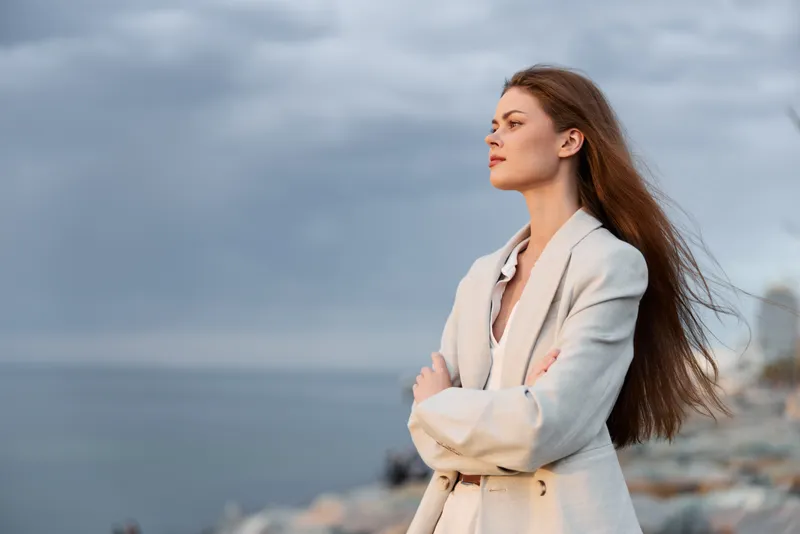 Woman in beige coat standing contemplatively by the sea with cloudy sky, exhibiting calm and thoughtful mood, nature background, autumn fashion style