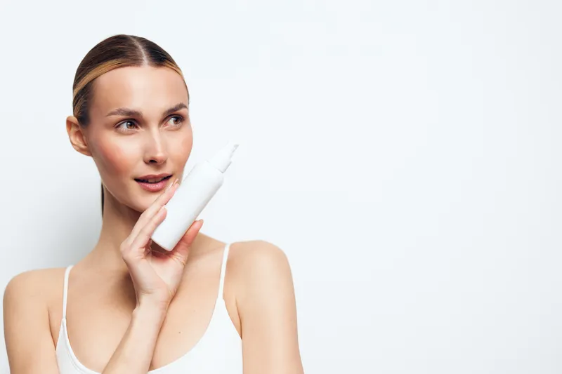 Young woman applying cream from a white bottle in a clean studio, presenting a skincare routine with soft lighting that creates a calm, fresh mood.