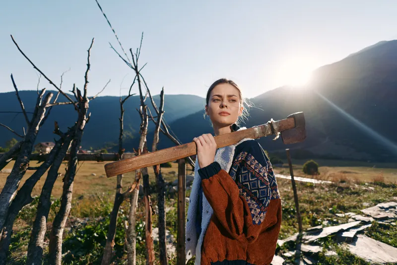 Woman carrying an axe on her shoulder in countryside near a rustic fence with mountains and sunrise in background, wearing a patterned sweater and looking confident.