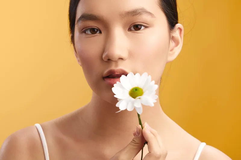 asian woman holds a white flower near her lips in a bright studio, her calm expression and gentle smile creating a fresh, serene mood suited for lifestyle and wellness themes, with grace and