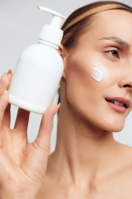 Young woman with clear skin applies moisturizer from a pump bottle onto her cheek, smiling softly, against a clean bright studio backdrop.