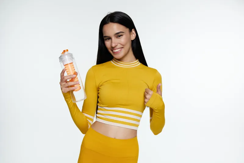Fit woman sport fitness wearing yellow activewear, holding a water bottle and smiling confidently against a clean white background.