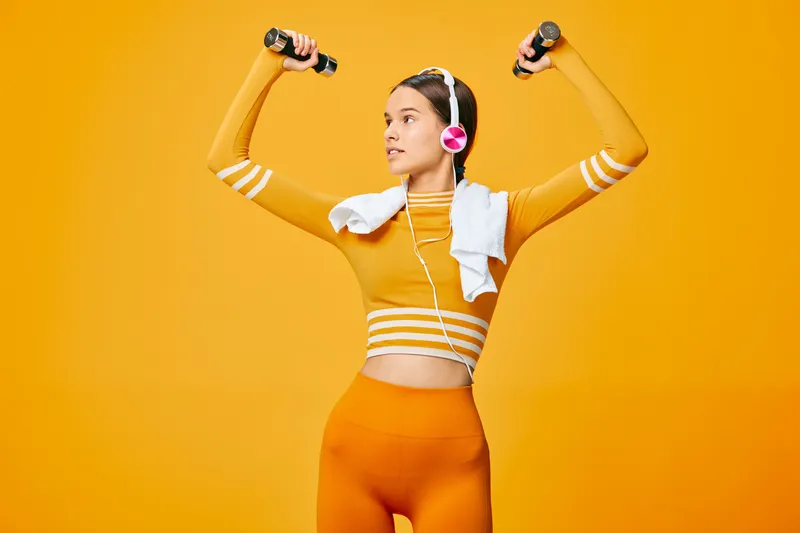 Dynamic woman in bright orange athletic wear lifts dumbbells with a towel and headphones, exuding fitness energy in a studio. Sport focused workout and health driven training vibe for active