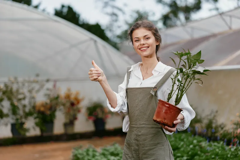 Smiling woman in green overalls holds a potted plant in a greenhouse, showcasing her passion for gardening and sustainable living