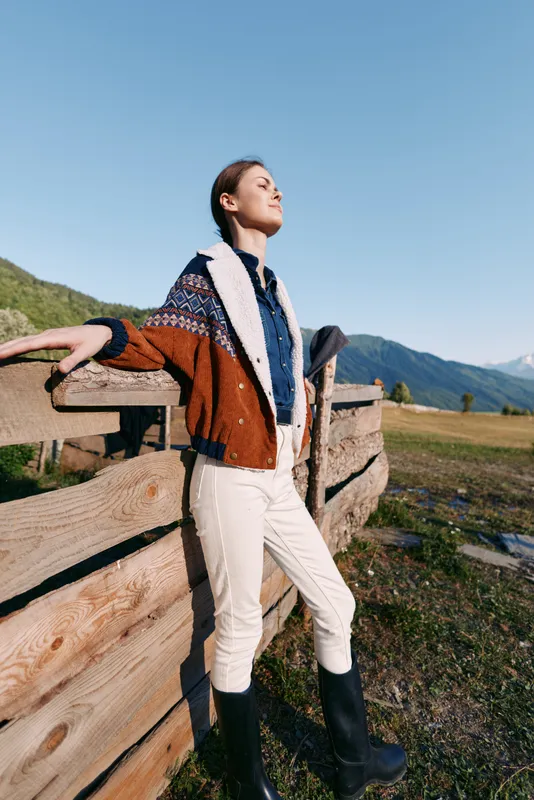woman in sweater and boots standing by wooden fence with mountains and countryside background, outdoors portrait evoking relaxed travel style, sunlight on scenic rustic landscape
