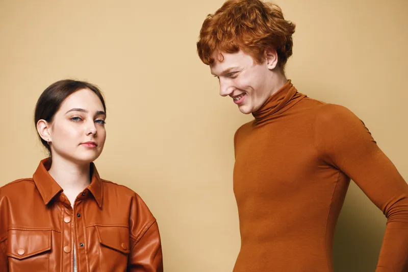 Young woman and man with red hair posing in stylish brown outfits against beige background. Fashion, casual style, portrait, looking at camera and smiling for modern clothing concept.