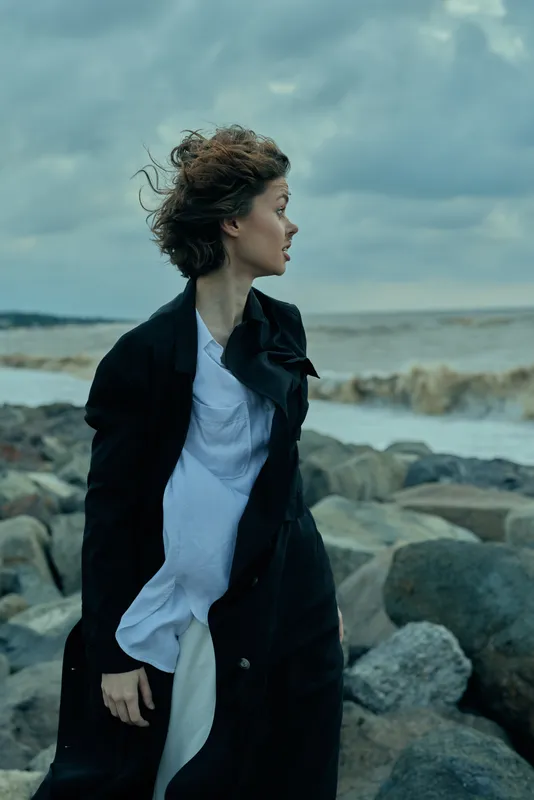 Windy coastal scene with a woman in a long coat standing among rocks as waves crash, creating a moody fashion moment by the sea