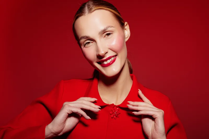 Smiling woman in red sweater poses against red background with bright makeup and glossy skin. Concept of fashion, beauty, confidence, and vibrant style in studio lighting.