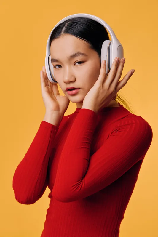 Young woman wearing white wireless headphones for music listening and enjoyment, posing in red sweater against orange background with focus on lifestyle and audio technology.