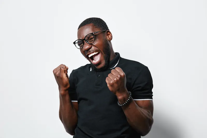 African American man with dark skin and glasses celebrating success with clenched fists and joyful expression in casual black polo shirt on white background.