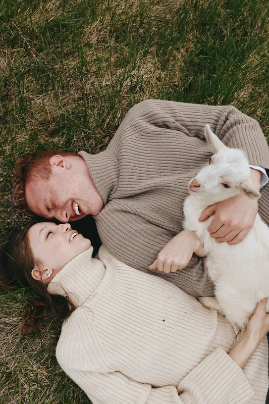 Cozy lifestyle photo of a couple lying on grass, embracing a white goat kid. Warm sweaters, natural light, intimate moment outdoors, playful bond, calm rural vibe and wholesome mood.