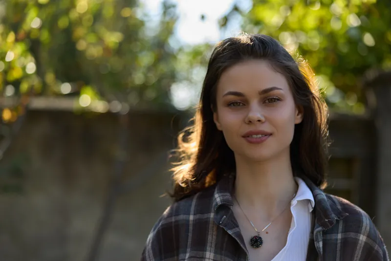 Smiling woman with long dark hair wearing a plaid shirt outdoors, casual lifestyle scene, natural light and greenery, portrait