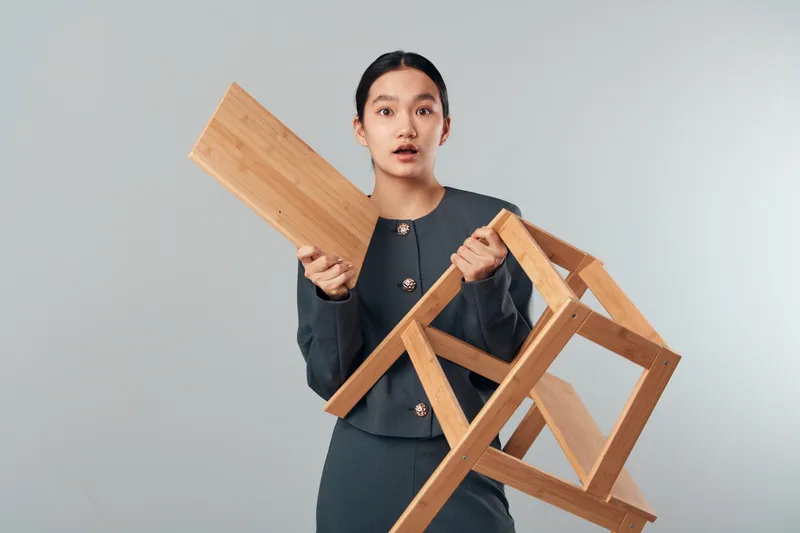 Woman holding wooden chair and plank in hands with surprised expression standing on light background. Female is wearing dark clothes and looking forward with open mouth and raised eyebrows.