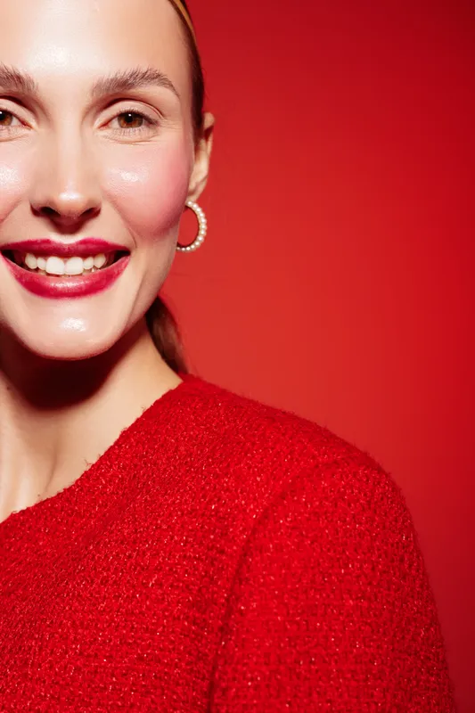 Smiling woman with red lipstick and hoop earrings wears red textured sweater against red background. Close-up portrait with natural skin glow and cheerful expression.