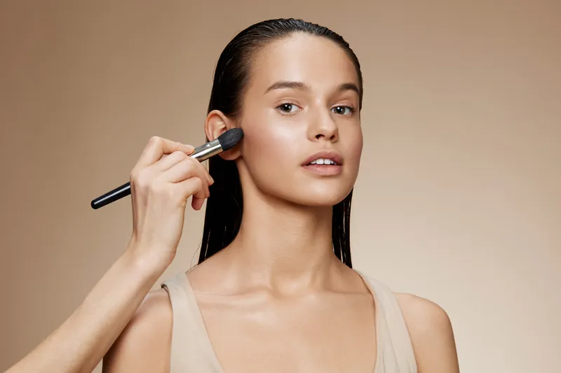 Young woman with natural makeup applying blush, showcasing a fresh look and soft lighting, ideal for beauty and skincare concepts