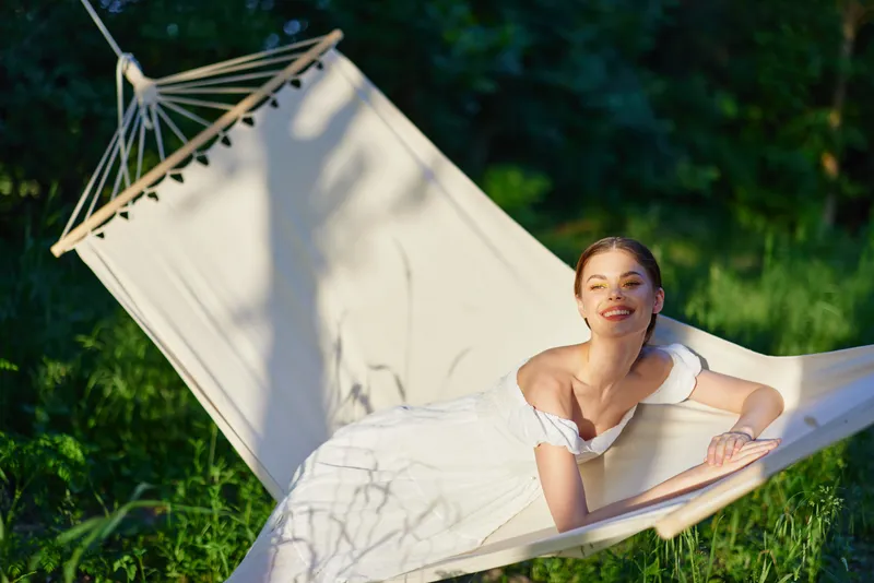 Young woman in a white dress relaxing in a hammock, smiling, surrounded by greenery on a sunny day, showcasing a serene summer atmosphere and joyful emotions