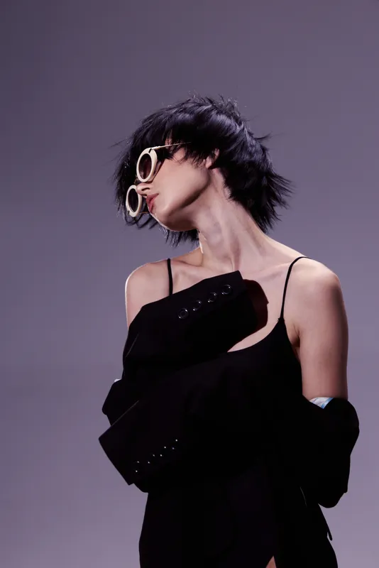 Stylish woman wearing sunglasses and black outfit posing with cool attitude in studio lighting.