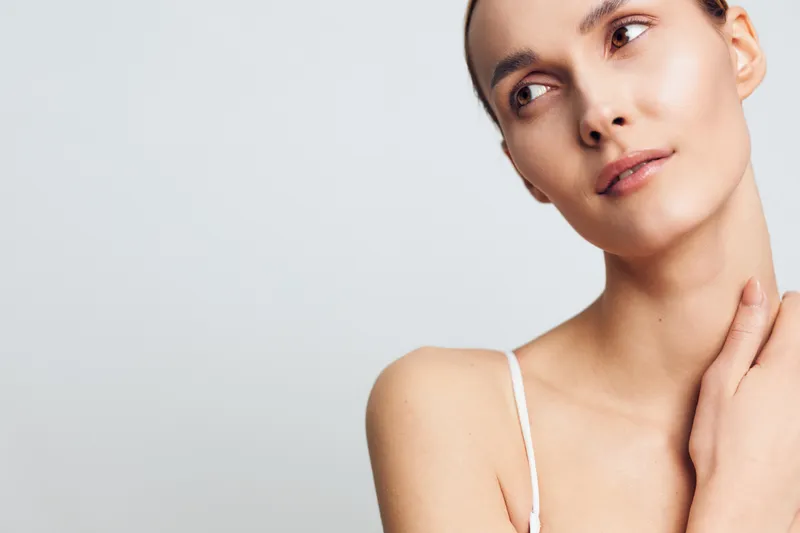 Portrait of a thoughtful young woman with clear skin looking away on a white background, wearing a white strap top, concept of beauty, skincare, and natural wellness.