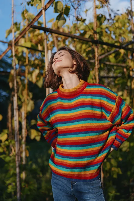Woman in colorful striped sweater stands outdoors in sunlight with eyes closed, portrait style, relaxed pose and hands on hips among garden vines, seasonal casual fashion.
