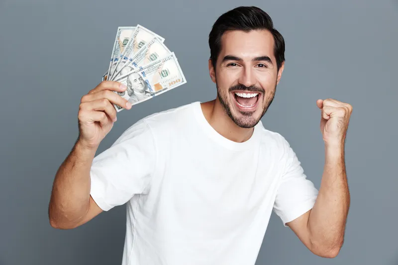 Happy smiling man celebrates success while holding cash in hand. He shows excitement and joy, dressed casually in a white t-shirt against a gray background.