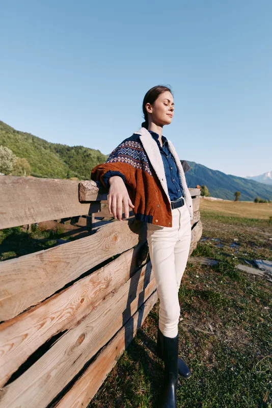 Woman leaning on wooden fence in mountains countryside, wearing patterned sweater and boots, standing in meadow under clear sky for relaxation, travel and outdoor lifestyle.