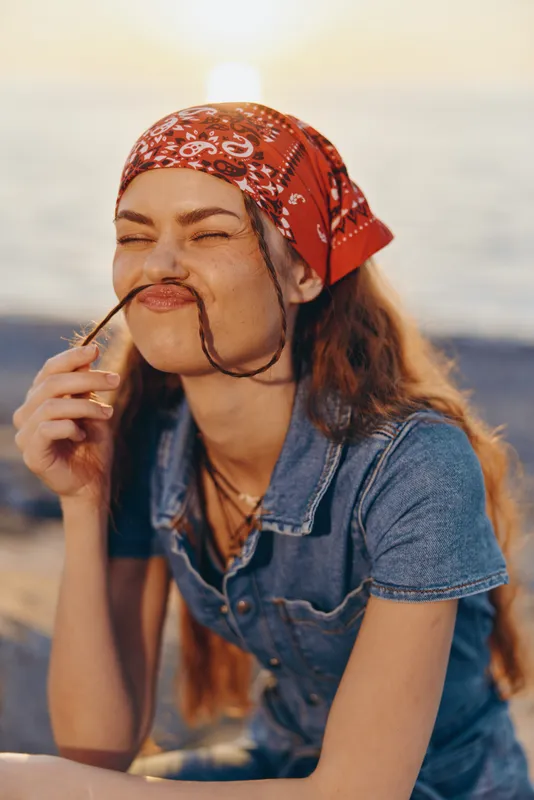 lifestyle woman with red bandana and denim shirt in modern boho-western street style, film color warm tones, playful mood near sunset by water
